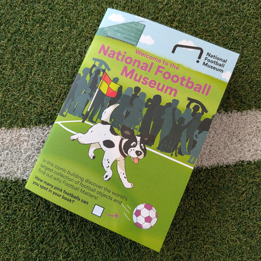 A printed activity book for the National Football Museum lies on green artificial turf with a white line. The cover shows a black-and-white dog on a soccer field with crowd silhouettes and the museum name in pink and white text.