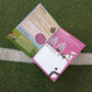 An open printed activity book laid on green artificial turf near a white line. The pages show soccer-themed graphics, text sections, and illustrations, including a pink page and a green page.