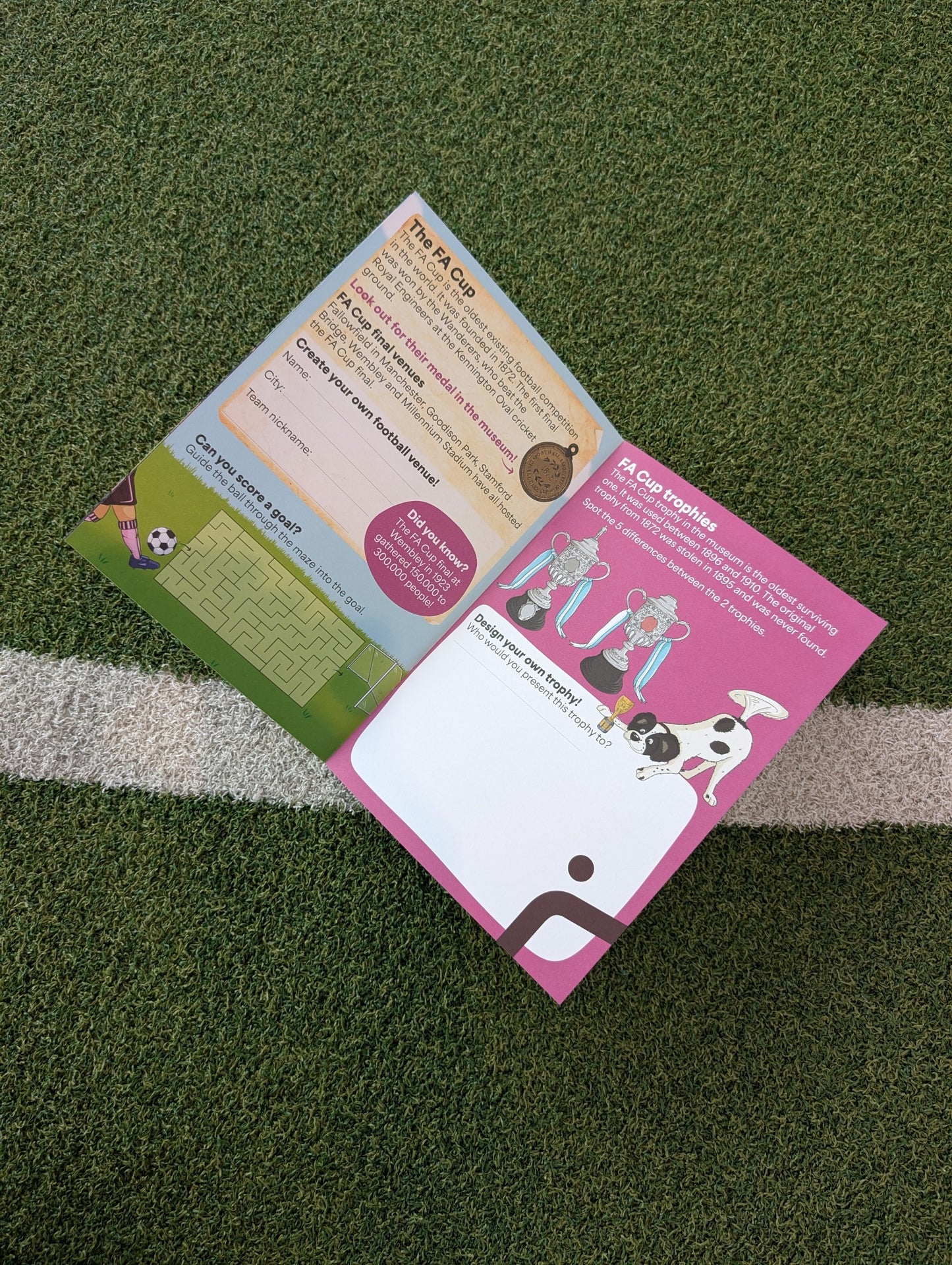 An open printed activity book laid on green artificial turf near a white line. The pages show soccer-themed graphics, text sections, and illustrations, including a pink page and a green page.