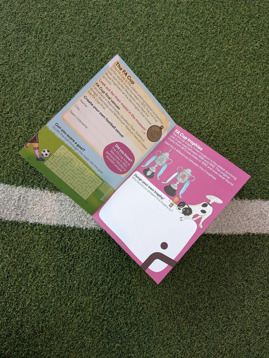 An open printed activity book laid on green artificial turf near a white line. The pages show soccer-themed graphics, text sections, and illustrations, including a pink page and a green page.